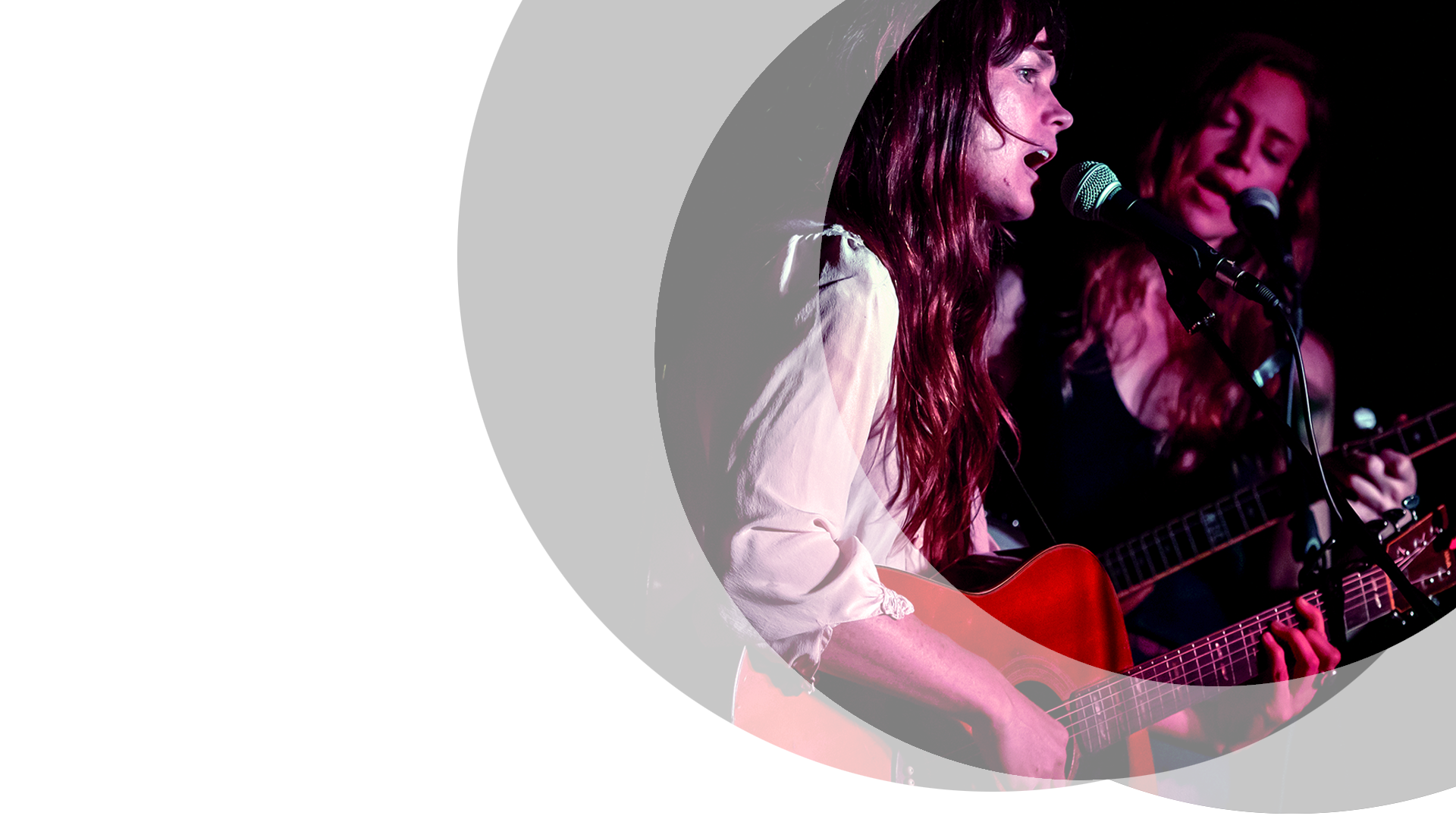 Two women with long hair singing into microphones set against a black background. The woman in the background is in low light and faces forward playing a bass guitar singing with her eyes closed. The woman in the foreground stands side on and is well lit. She is playing a deep red/brown acoustic guitar while singing.